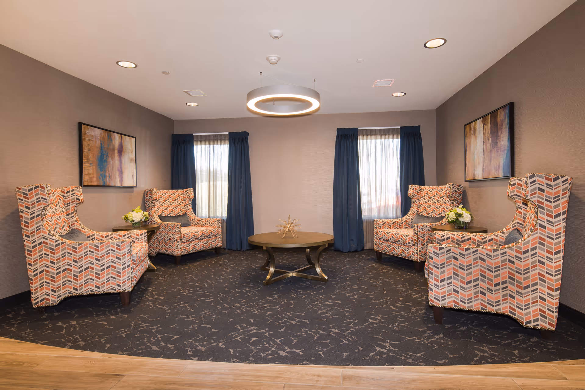 A cozy seating area in a senior living facility with four patterned armchairs arranged around a round wooden coffee table. The room has two windows with sheer curtains and dark blue drapes, two abstract paintings on the walls, and a modern circular ceiling light fixture. Small side tables with flower arrangements are placed between the chairs.