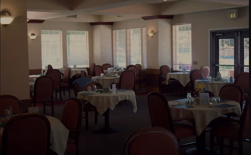 Interior view of a dining room in a senior living facility with multiple round tables covered with white tablecloths, each set with glasses, napkins, and condiments. The room has several windows with blinds allowing natural light to enter, and upholstered chairs are arranged around the tables.