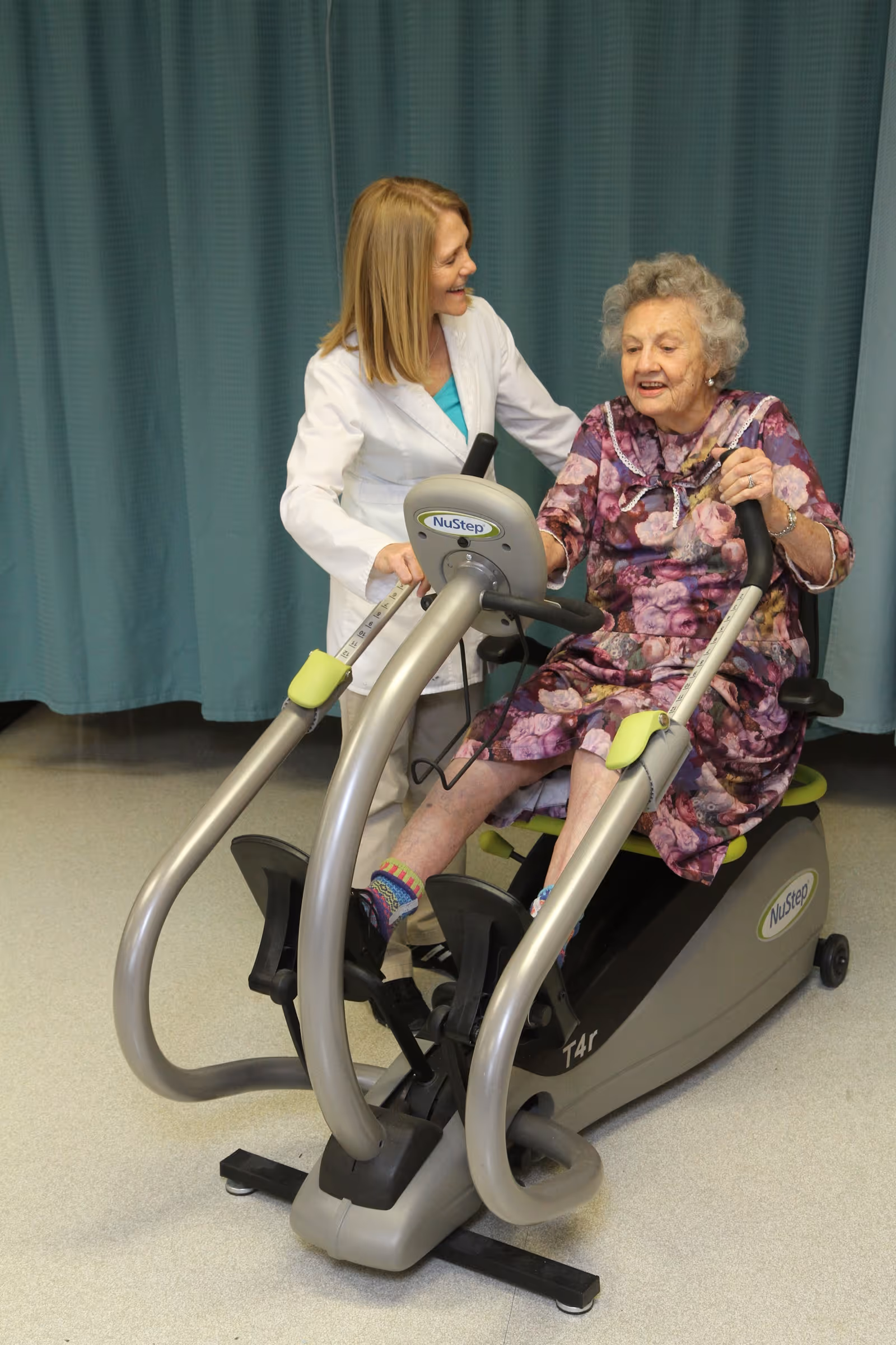 An elderly woman in a floral dress is seated on a NuStep exercise machine, pedaling with her feet while holding the handles. A healthcare professional in a white coat stands beside her, smiling and providing support. The background features a teal curtain and a light-colored floor.