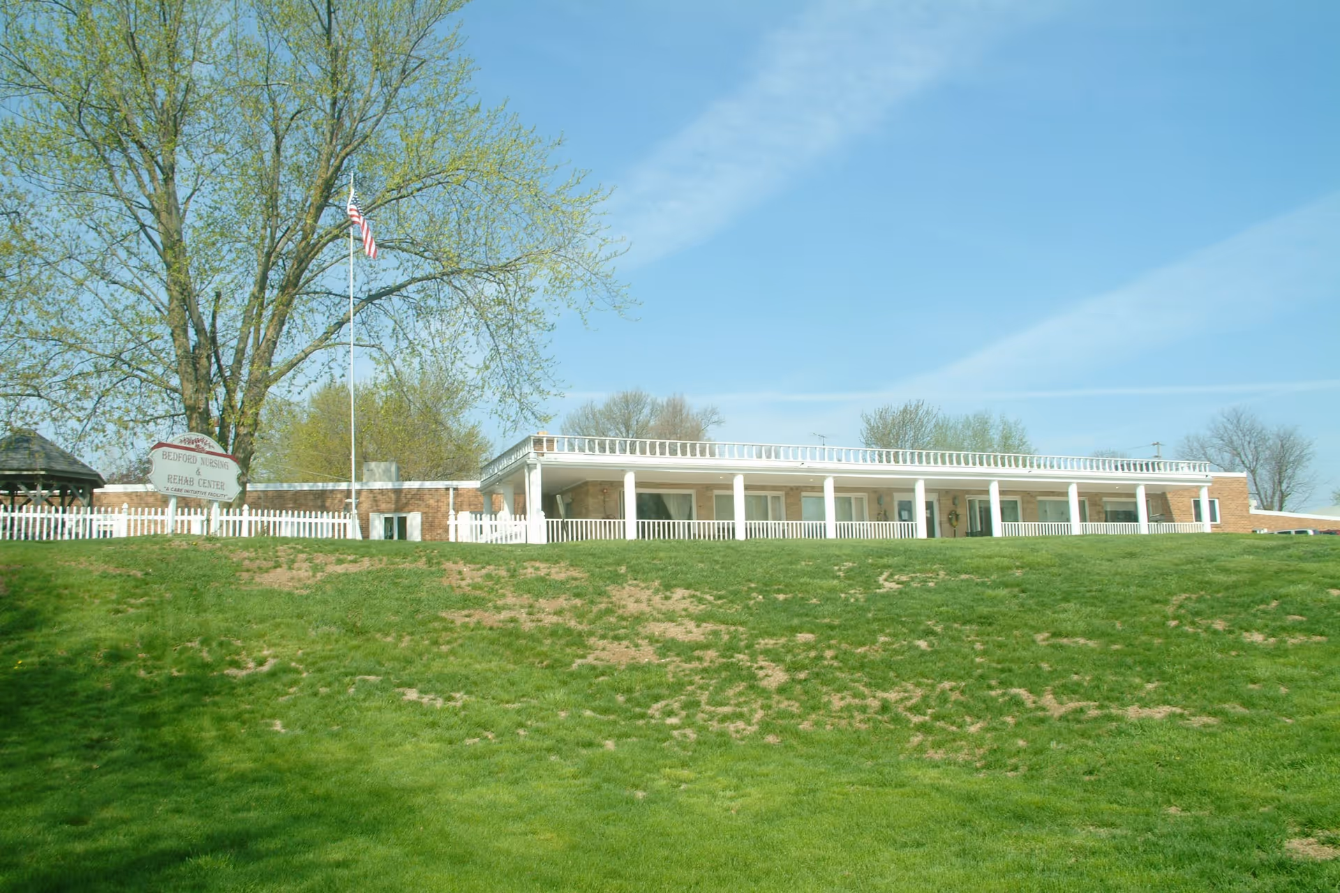 Exterior view of Bedford Specialty Care Rehab Center building with a white picket fence, a flagpole with an American flag, and a large tree on a grassy hill under a clear blue sky.