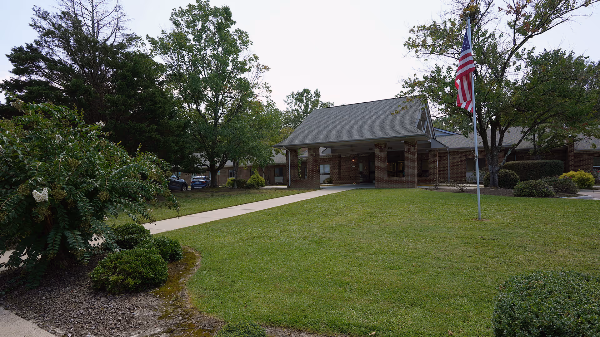 Front exterior view of Pinehurst Healthcare & Rehabilitation Center showing a brick building with a covered entrance, a walkway leading to the entrance, a flagpole with an American flag, and surrounding green lawn and trees.