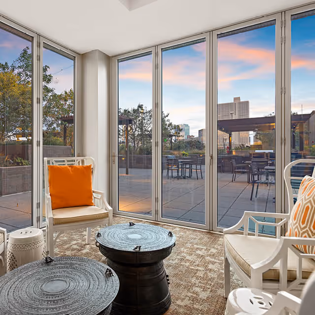 A bright indoor seating area with large floor-to-ceiling glass doors opening to an outdoor patio. The room features white chairs with orange and patterned cushions, two round black tables, and a beige carpet. Outside, there are tables and chairs on the patio with trees and city buildings visible in the background under a colorful sky at sunset.