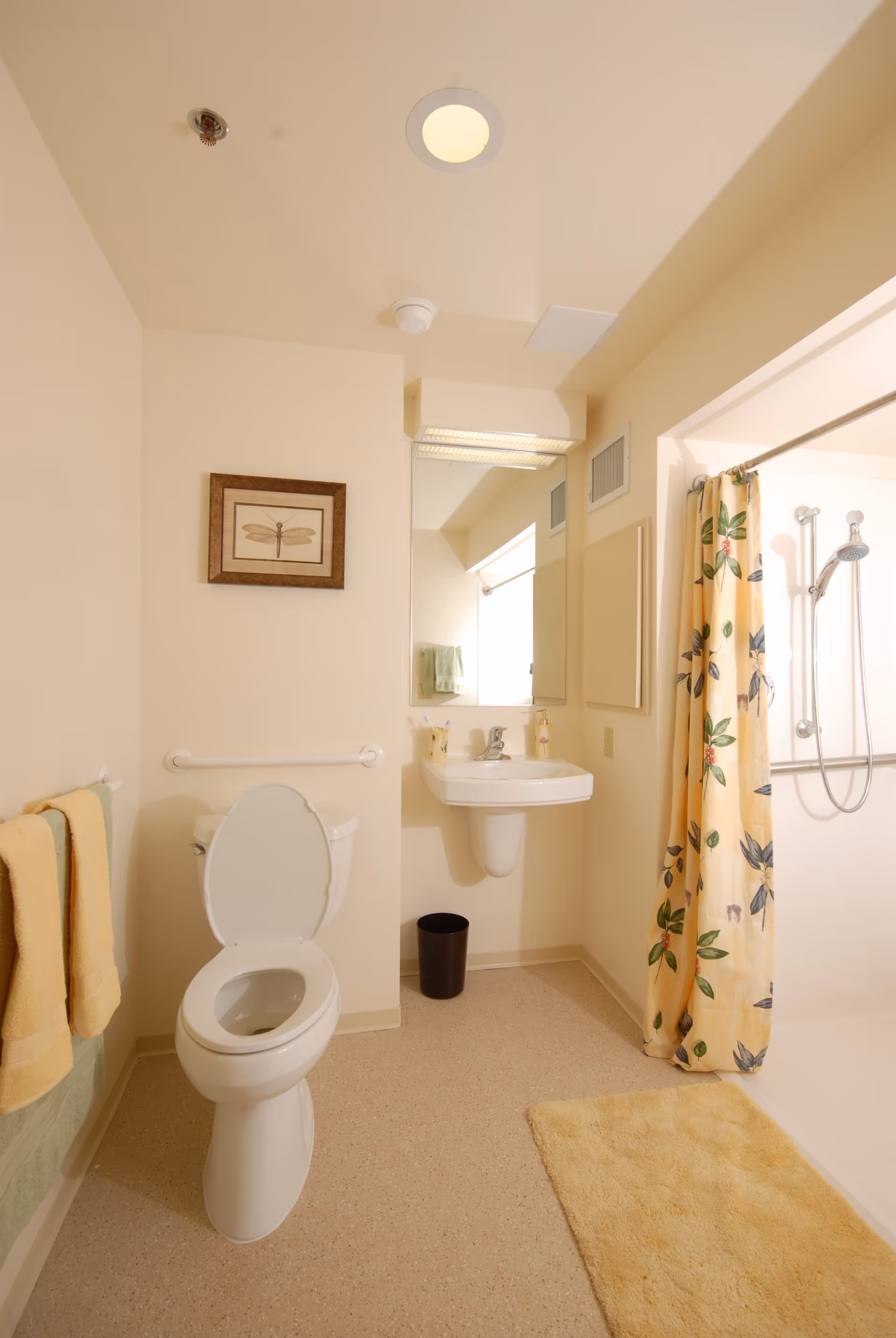 A clean, accessible bathroom with toilet, wall-mounted sink, mirror, and floral-curtain shower.