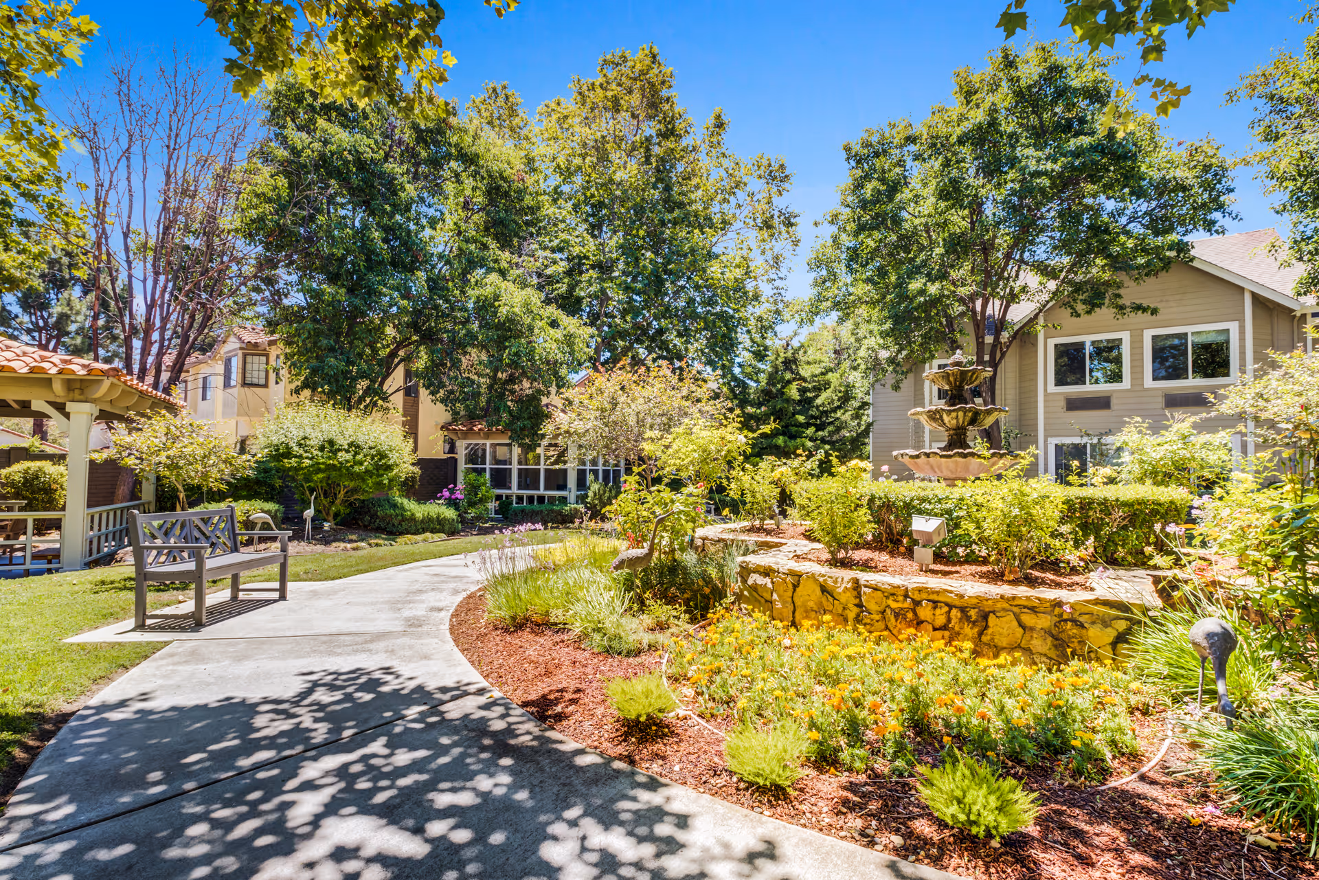 Sunlit landscaped courtyard with a winding paved path, bench, flower beds, and a multi-tiered fountain in front of residential buildings.