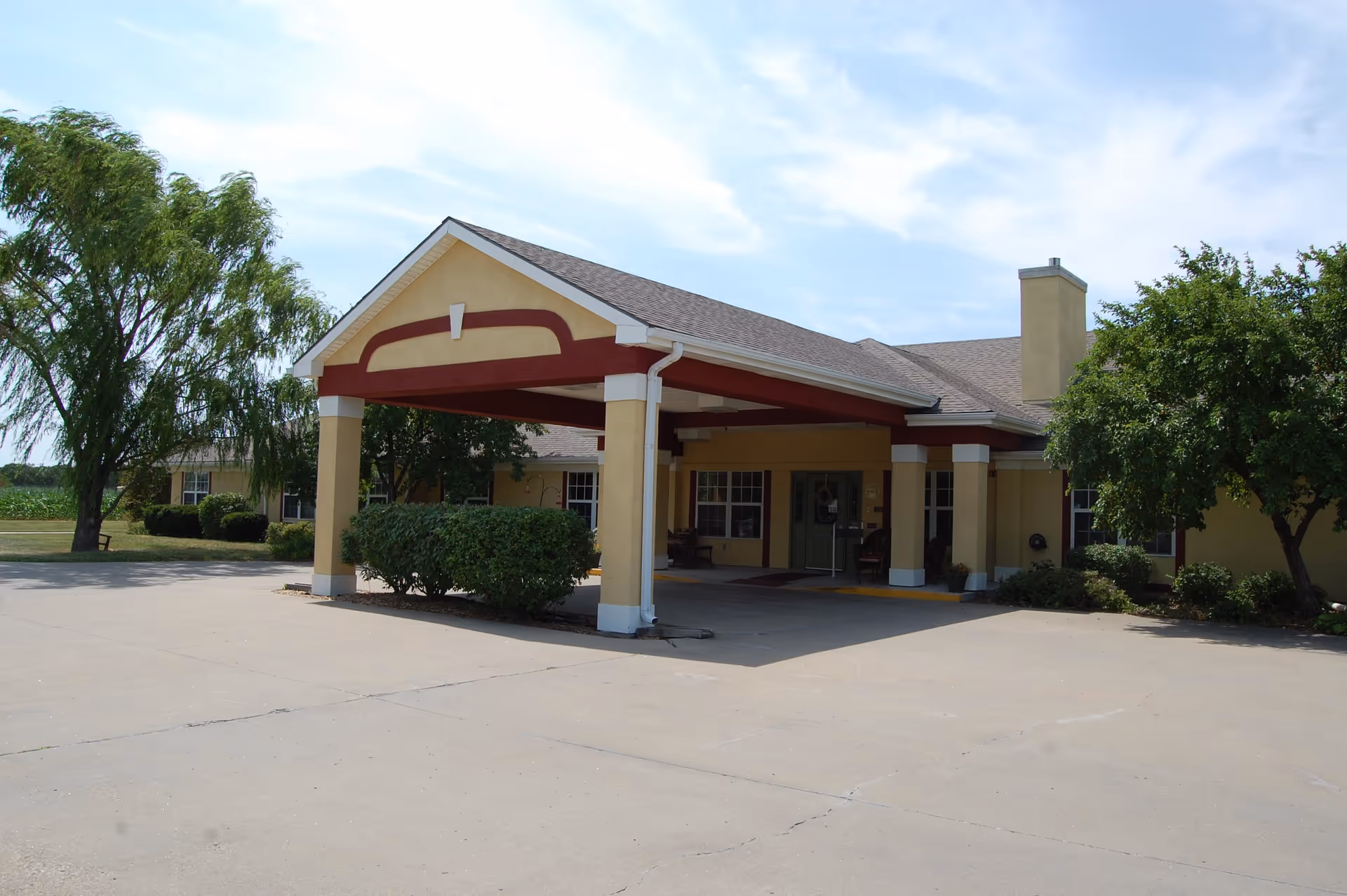 Exterior view of the entrance to Vintage Park at Osage City, showing a covered driveway with yellow and red pillars, surrounded by trees and bushes under a partly cloudy sky.