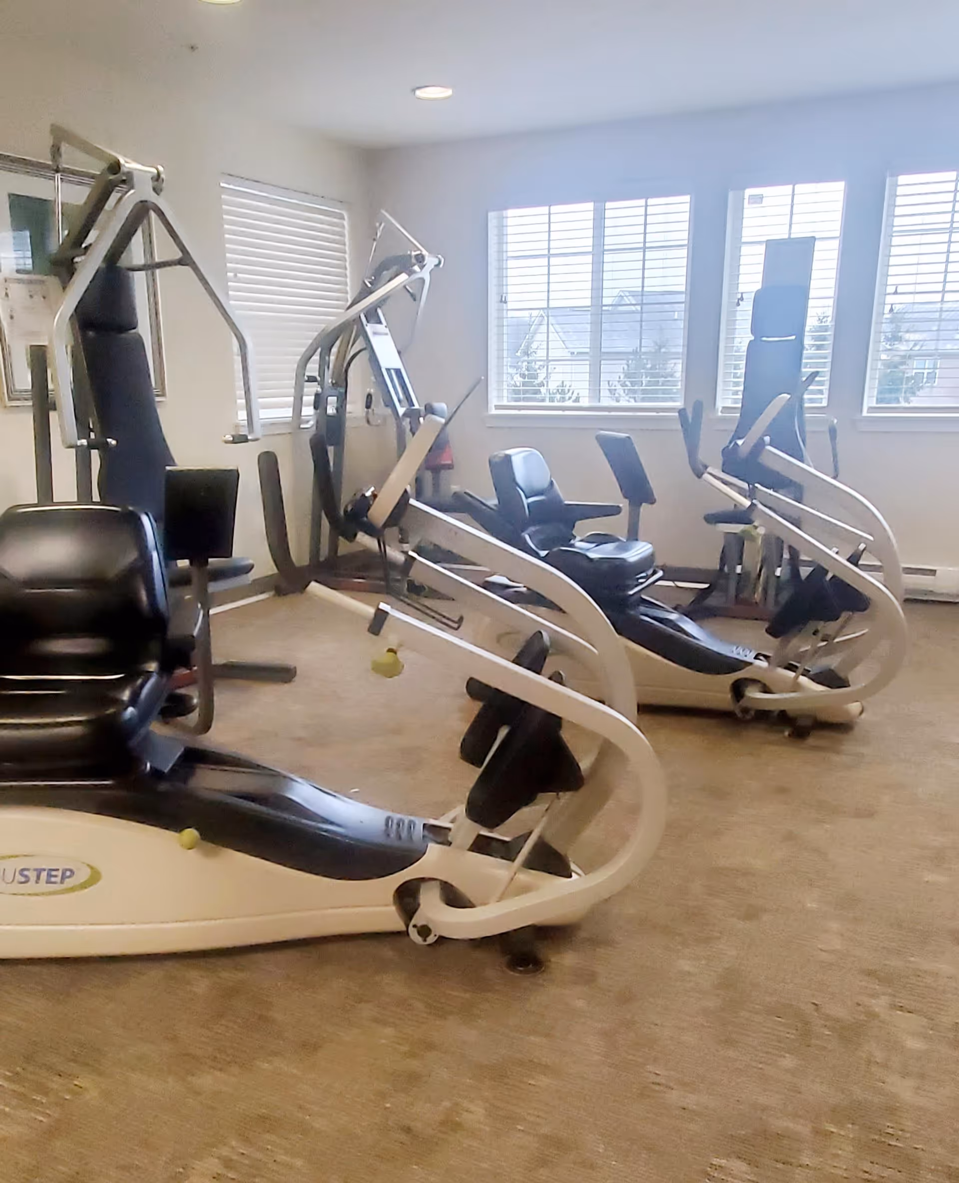 A small exercise room with several pieces of fitness equipment including recumbent bikes and weight machines. The room has beige carpet, white walls, and windows with white blinds letting in natural light.