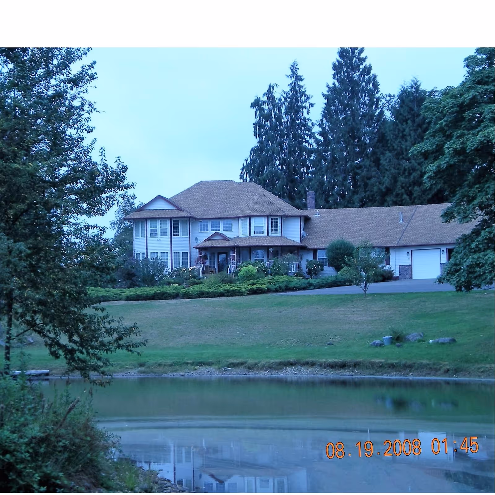 A large two-story house with a brown roof and white exterior, surrounded by trees and greenery. In the foreground, there is a pond reflecting the house and the surrounding landscape. The sky is clear and the scene appears peaceful and serene.