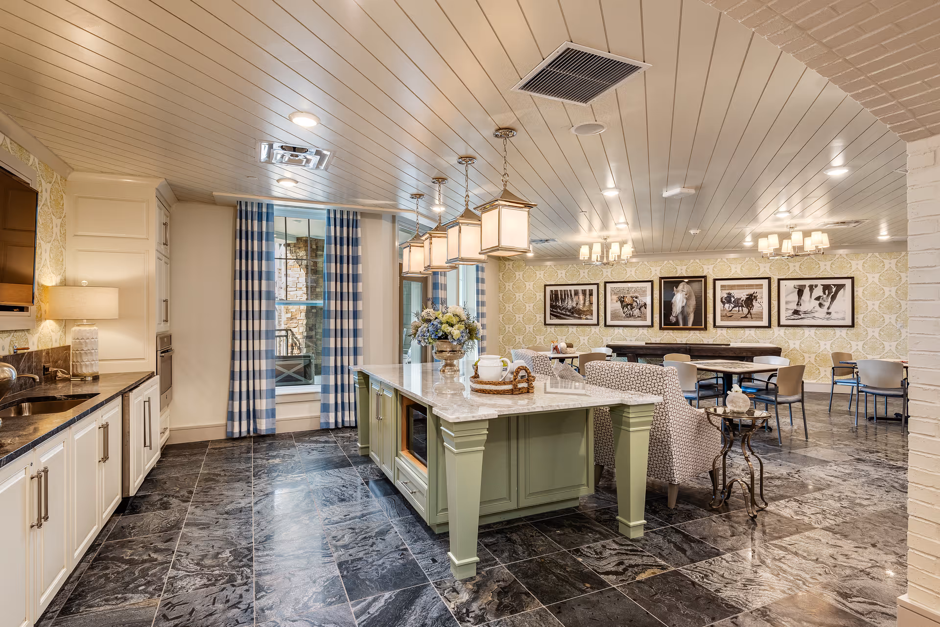 A spacious, well-lit interior room featuring a kitchen area with white cabinetry, a marble countertop island with green base, and a seating area with patterned armchairs. The room has large windows with blue and white checkered curtains, dark tiled flooring, and framed horse-themed artwork on the wallpapered wall in the background. Multiple pendant lights hang above the island, and there are tables and chairs arranged for dining or socializing.