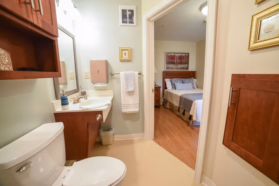View of a bathroom with a toilet, sink, mirror, and wooden cabinets. A towel hangs on a rack next to the sink. Through an open doorway, a bedroom with a bed, nightstand, and framed artwork on the wall is visible.