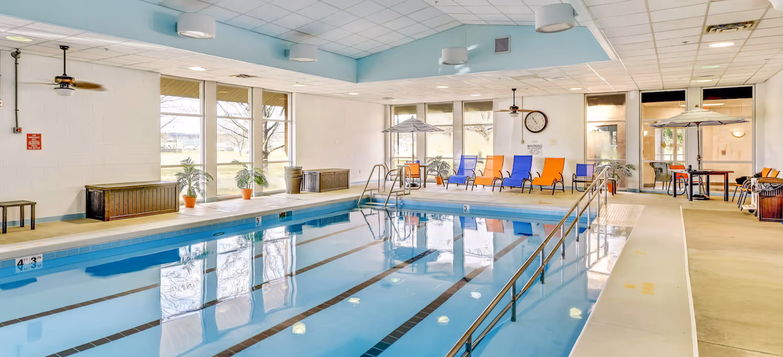 Indoor swimming pool area with lounge chairs, umbrellas, handrails and large windows letting in daylight.