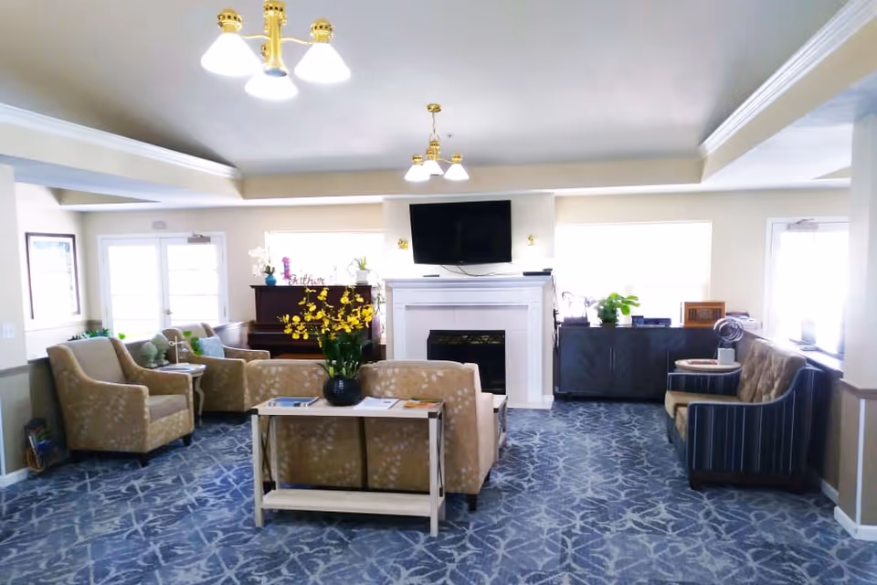 A bright and spacious senior living facility common area with patterned blue carpet, beige and striped armchairs, a beige sofa, a wooden table with a vase of yellow flowers, a piano, a fireplace with a mounted flat-screen TV above it, and large windows letting in natural light.