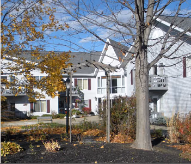 Outdoor view of a senior living facility with white buildings, maroon shutters, and a courtyard featuring a pergola, trees with autumn leaves, and landscaped garden beds.