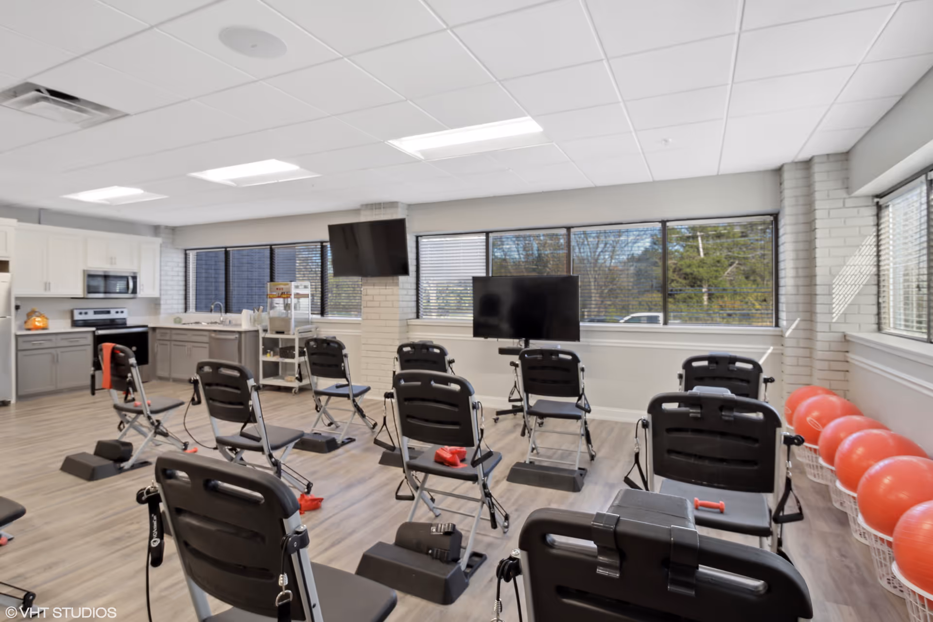 A bright, spacious exercise room with multiple black exercise chairs arranged in rows facing two large flat-screen TVs mounted on the wall. The room has large windows letting in natural light, a kitchen area with cabinets, a microwave, and an oven on the left side, and a row of red exercise balls stored in baskets on the right side.