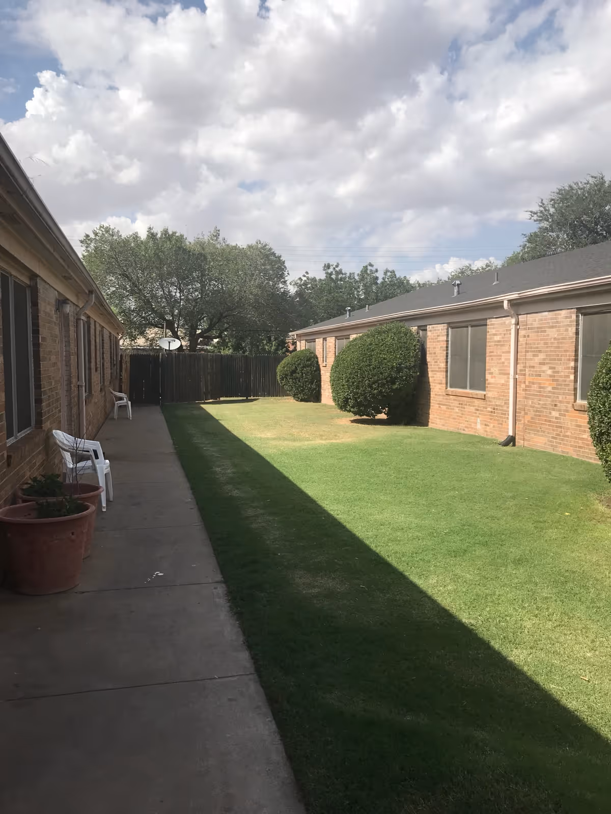 Outdoor view of a grassy courtyard area between two single-story brick buildings with windows. There are neatly trimmed bushes along the building on the right and a concrete walkway on the left with white plastic chairs and large potted plants. The sky is partly cloudy.