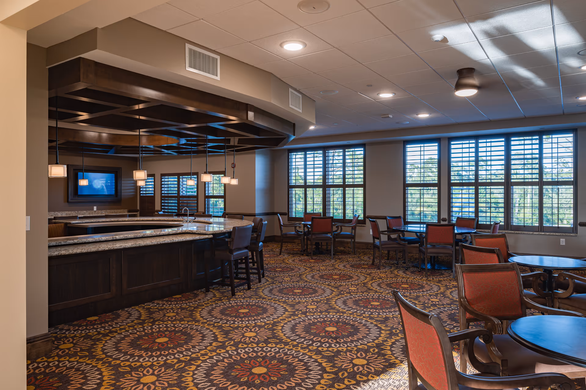 A spacious dining area in Palagio Senior Living featuring multiple round tables with red cushioned chairs. The room has large windows with shutters allowing natural light to fill the space. There is a bar counter with granite countertops and several hanging pendant lights above it. The carpet has a colorful circular pattern, and the ceiling has recessed lighting.