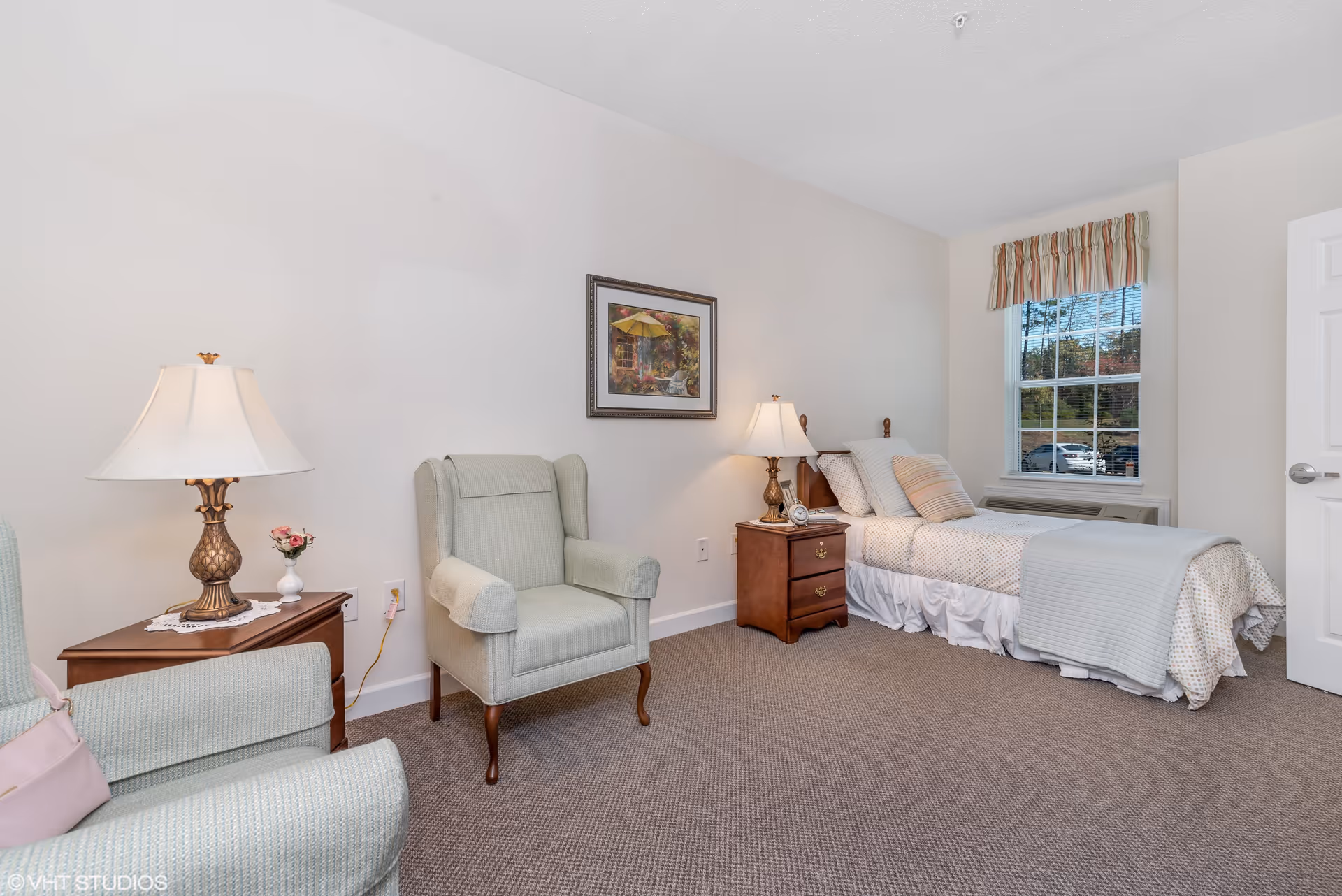 A cozy bedroom in a senior living facility featuring a single bed with patterned bedding and pillows, a wooden nightstand with a lamp and clock, two upholstered armchairs, a wooden side table with a lamp and small flower vase, a window with striped valance, and a framed painting on the wall.