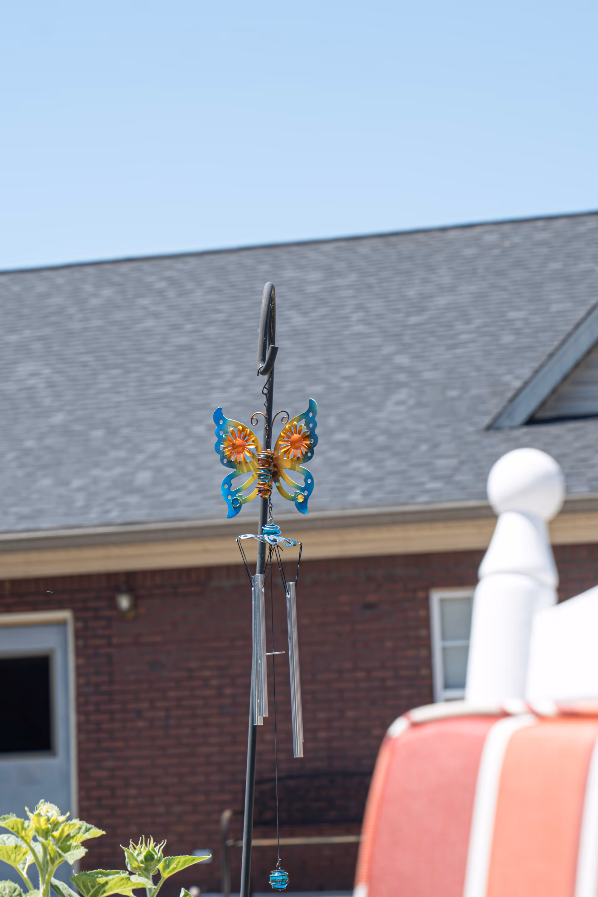 A colorful butterfly-shaped wind chime hanging outdoors with a brick building and a gray shingled roof in the background. Part of a red and white outdoor chair is visible in the foreground.