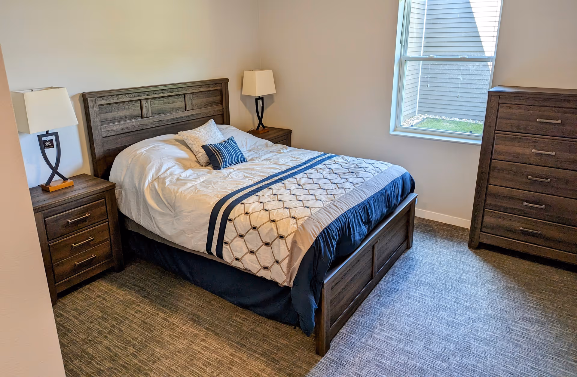 A bedroom with a wooden bed frame and headboard, a neatly made bed with white and blue bedding, two decorative pillows, two matching wooden nightstands each with a lamp, a wooden dresser, and a window letting in natural light.