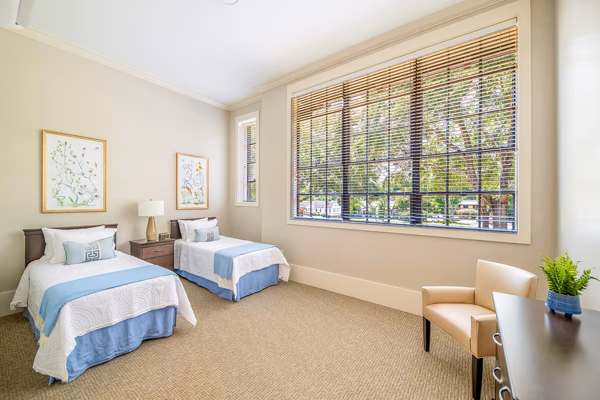 A bright bedroom with two twin beds dressed in white and light blue bedding. Between the beds is a dark wooden nightstand with a lamp and a small clock. Above each bed hangs a framed floral artwork. A large window with wooden blinds lets in natural light, and a beige armchair sits next to a dark wooden dresser with a small green plant on top. The walls are painted light beige and the floor is carpeted.