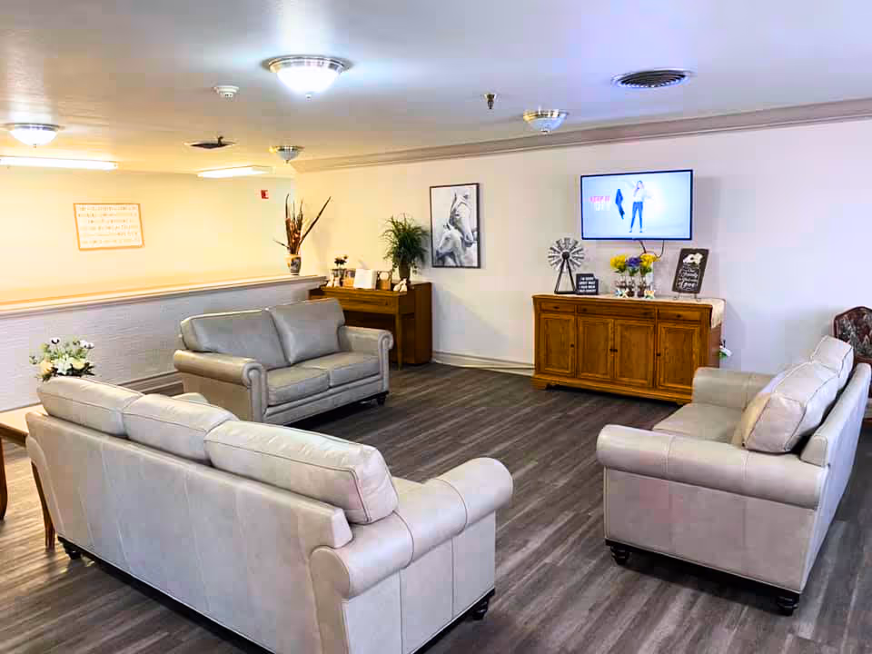 A cozy living room area with three light gray leather sofas arranged around a wooden cabinet with a TV mounted on the wall above it. The room has wood flooring, white walls, ceiling lights, and decorative items including framed pictures, plants, and flowers.