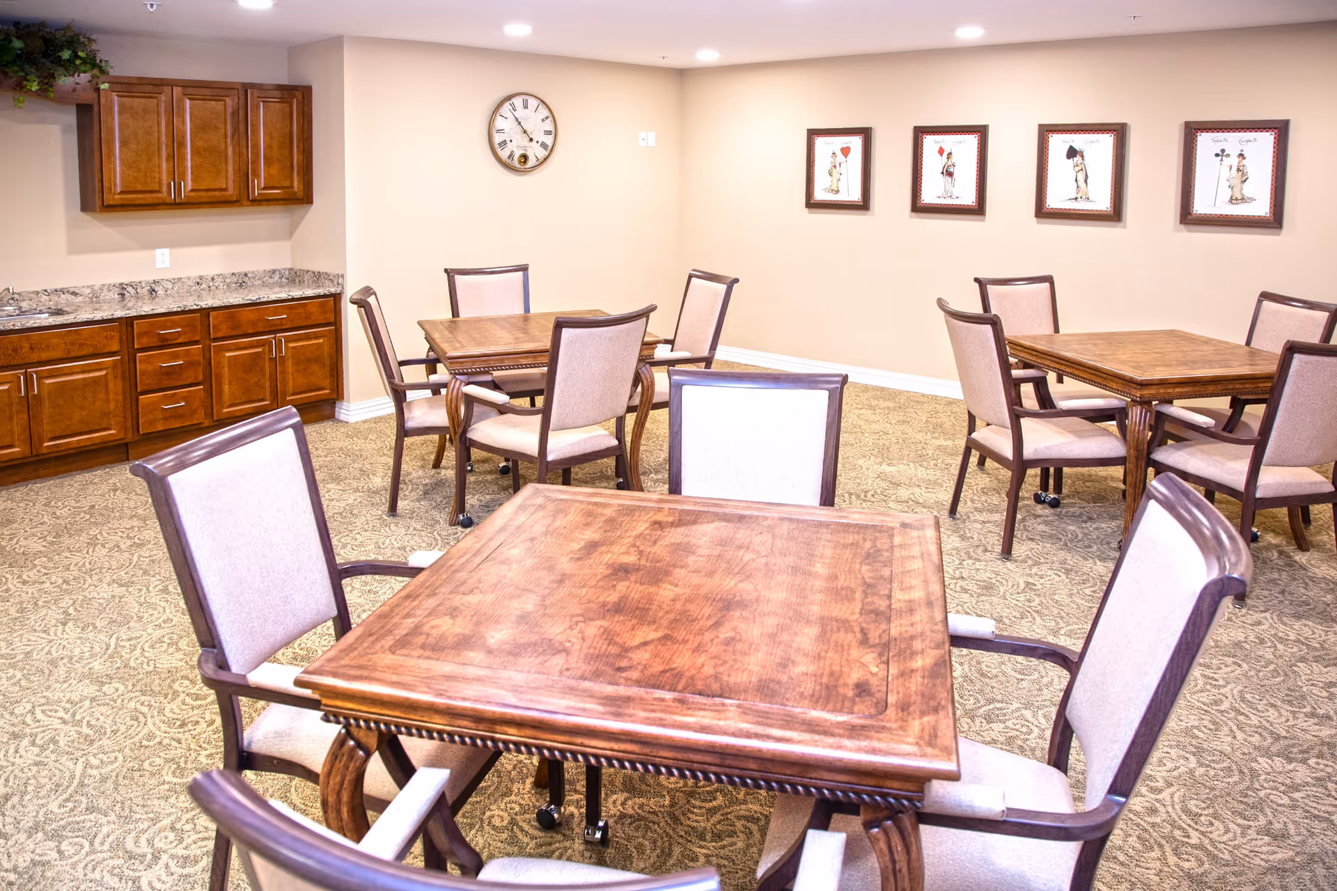 A well-lit room with multiple wooden tables and cushioned chairs arranged around them. The walls are beige with four framed pictures of playing cards and a round clock. There is a wooden cabinet with a granite countertop and a small sink on the left side of the room. The floor is carpeted with a patterned design.