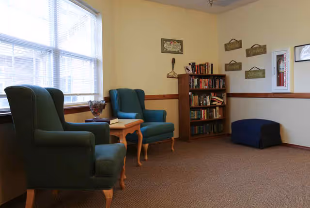 Small seating area with two green armchairs, a wooden side table, bookshelf and window in a senior living facility.