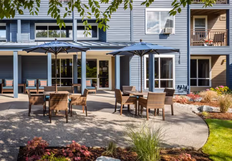 Outdoor patio area at Cogir at The Narrows with several tables and chairs under large umbrellas. The patio is adjacent to a building with blue and beige siding, large windows, and a balcony. There are plants and landscaping around the patio area.