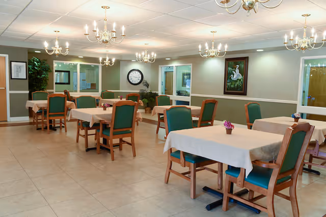 A dining room with several tables covered with beige tablecloths and green cushioned wooden chairs. The room has tiled floors, beige and green walls, multiple chandeliers hanging from the ceiling, a large clock on the far wall, a painting of white birds, and some potted plants.