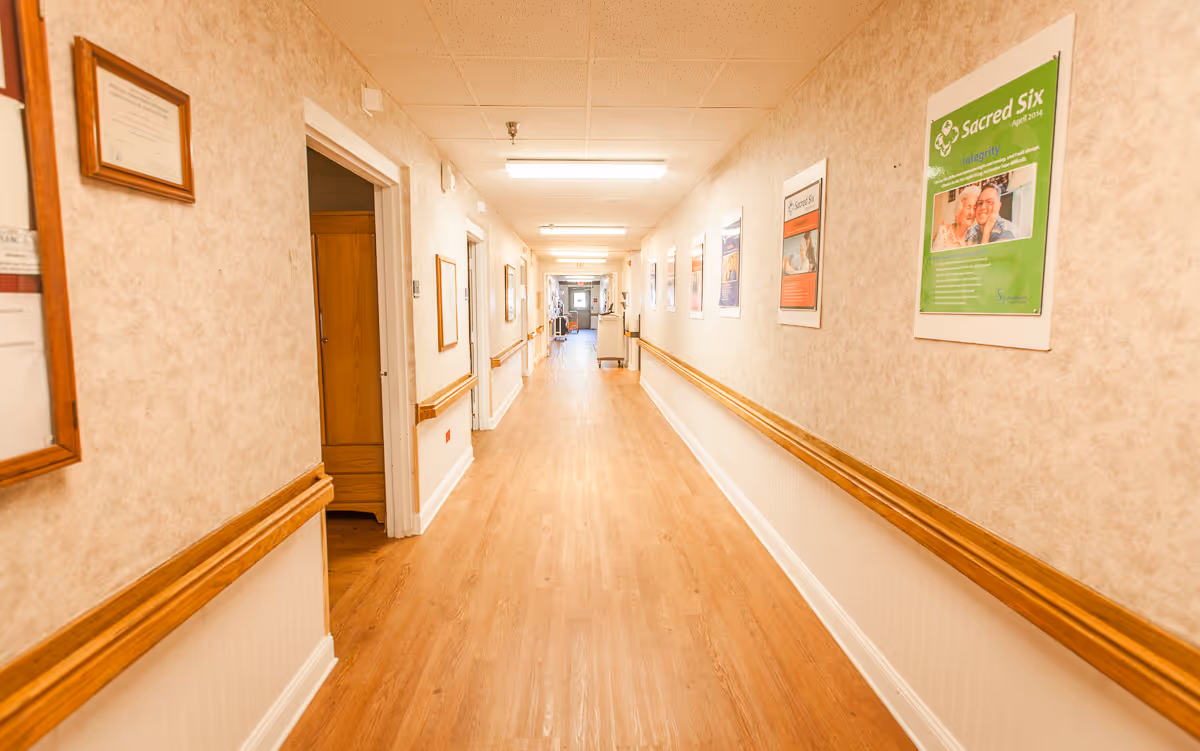 A long, well-lit hallway in a senior living facility with wooden handrails on both sides and framed posters and certificates hanging on the walls. The floor is made of light wood, and there are several doorways along the hallway.