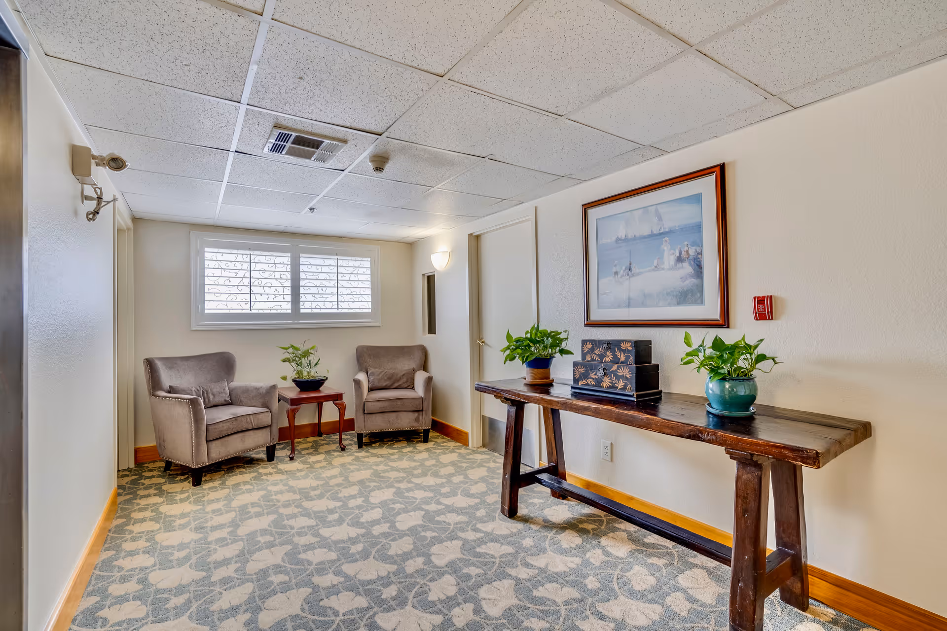 Small seating area in an assisted living facility with two armchairs by a window and a long wooden console table topped with potted plants and framed artwork.