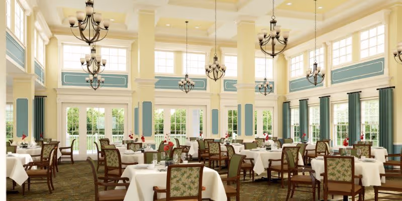 A spacious and elegant dining room with high ceilings, large windows, and multiple chandeliers. The room is furnished with round tables covered with white tablecloths and surrounded by wooden chairs with patterned cushions. The walls are painted in soft yellow and blue tones, and the windows have green curtains. The room is bright with natural light coming through the windows.