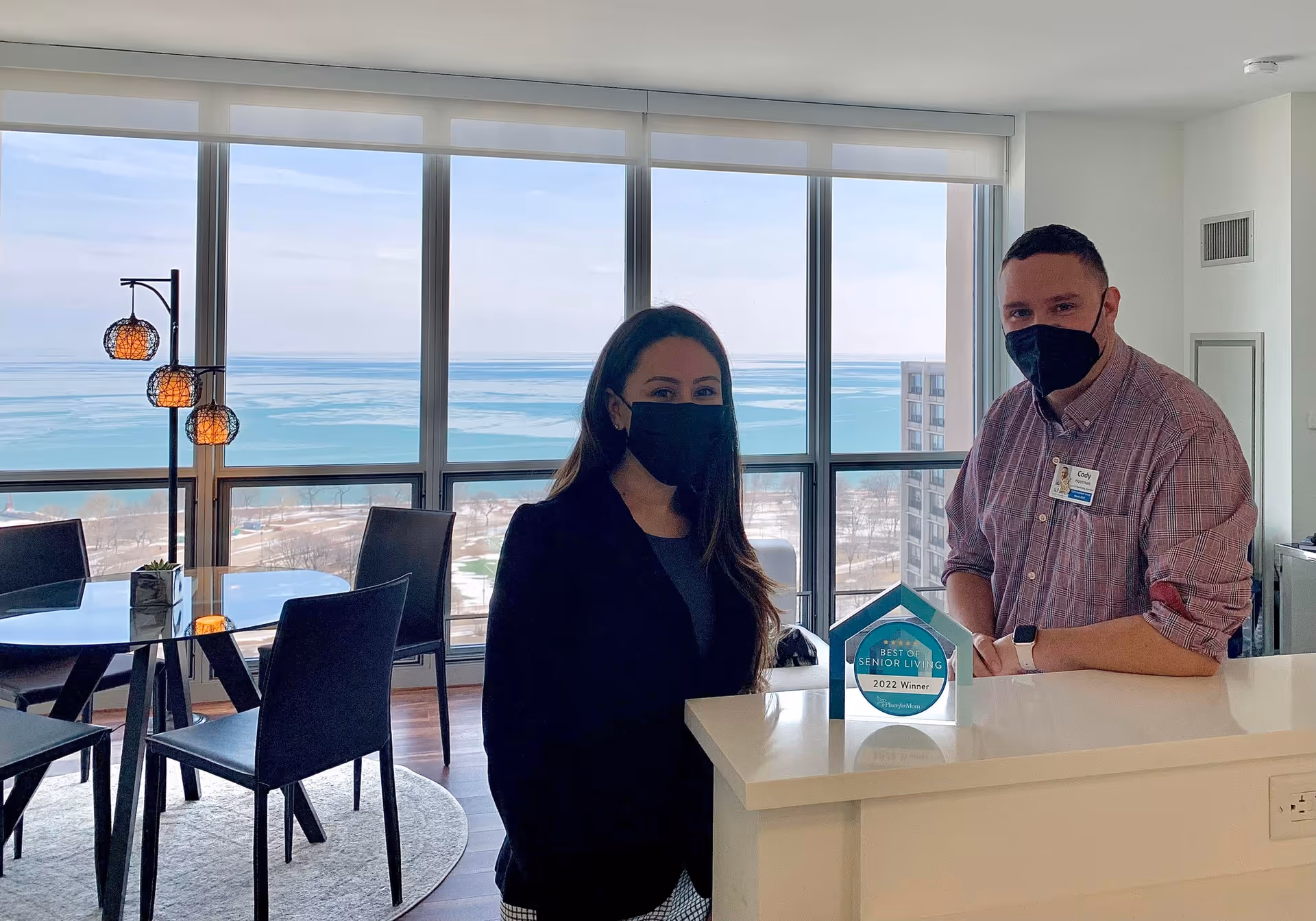 Two people wearing black face masks standing inside a modern room with large windows overlooking a body of water. One person is standing behind a white counter with a 'Best of Senior Living 2022 Winner' award displayed on it. The room has a dining table with chairs and a floor lamp with three spherical lights.