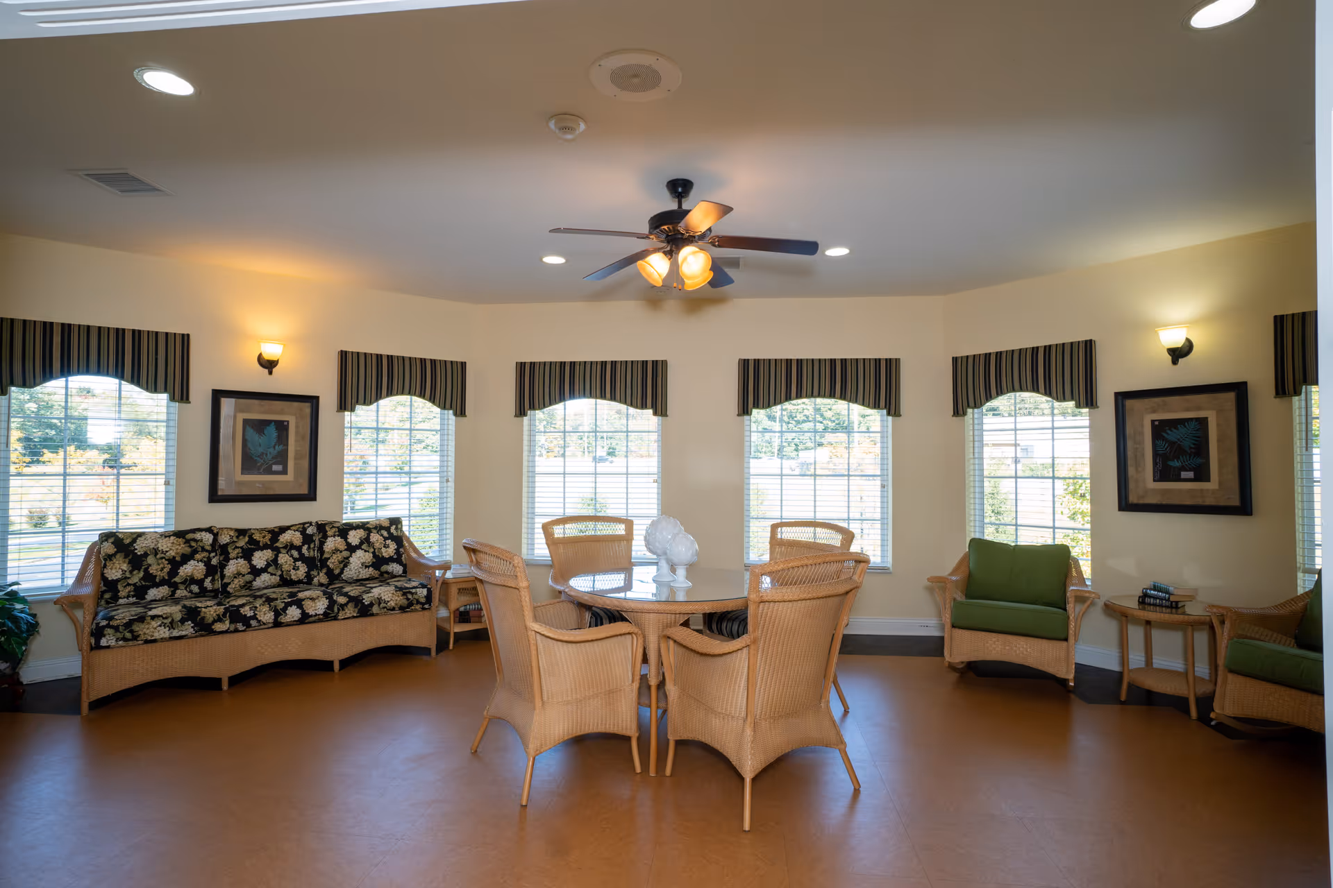 A bright and cozy common area with a round glass table surrounded by six wicker chairs. There is a floral-patterned wicker sofa on the left and two green cushioned wicker armchairs on the right. The room has five large windows with striped valances, framed artwork on the walls, and a ceiling fan with lights.