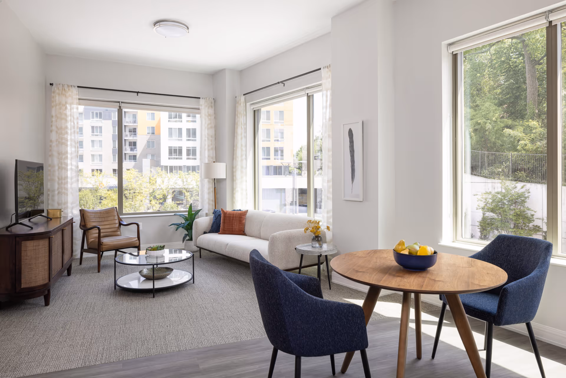 A bright and modern living room with large windows showing views of nearby buildings and greenery. The room features a white sofa with colorful pillows, a wooden armchair, a round glass coffee table, a wooden TV stand with a flat-screen TV, and a small round dining table with two blue upholstered chairs. The walls are white, and the floor is a combination of carpet and wood. Light curtains frame the windows, and a small side table with a vase of flowers is next to the sofa.