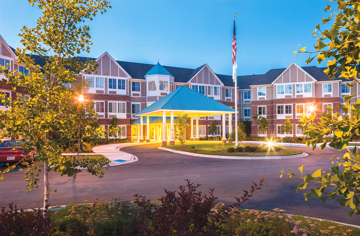 Front entrance of The Rivers Grosse Pointe senior living building with a covered porte-cochere, flagpole, and landscaped circular driveway at dusk.