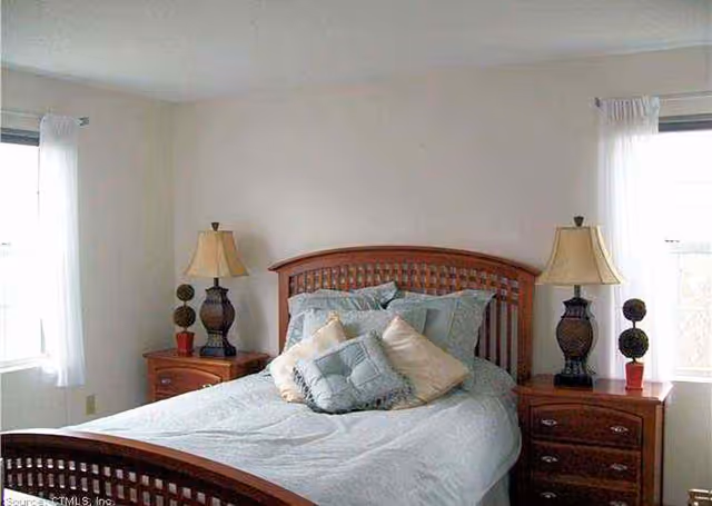 Simple bedroom with a wooden bedframe, light blue bedding, two matching bedside tables with lamps and windows on either side.