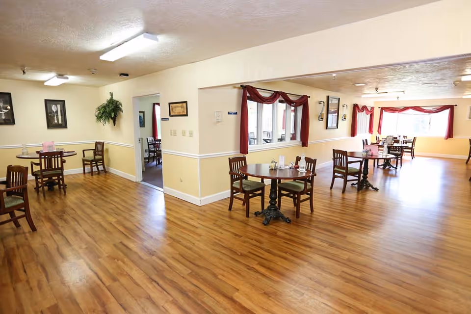 A spacious dining room with wooden floors and several round wooden tables, each surrounded by four chairs with green cushions. The walls are painted in a light yellow color with white trim, and there are framed pictures and a hanging plant on the walls. Large windows with red curtains allow natural light to fill the room.