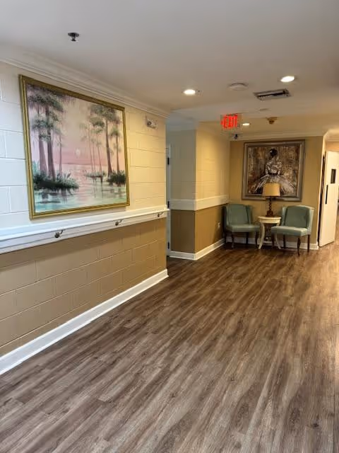 Well-lit interior hallway with wood-look flooring, handrails, framed paintings, and two chairs beside a small table beneath an exit sign.