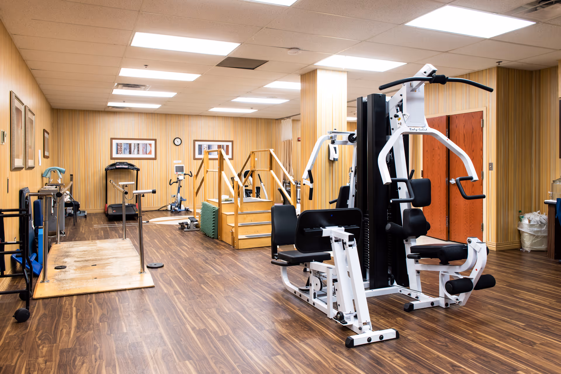 A fitness and rehabilitation room with various exercise equipment including a multi-station gym machine, stationary bikes, a treadmill, and a wooden ramp with handrails. The room has wood-patterned flooring, beige striped wallpaper, and fluorescent ceiling lights. There are framed pictures on the walls and a clock above the exercise machines.