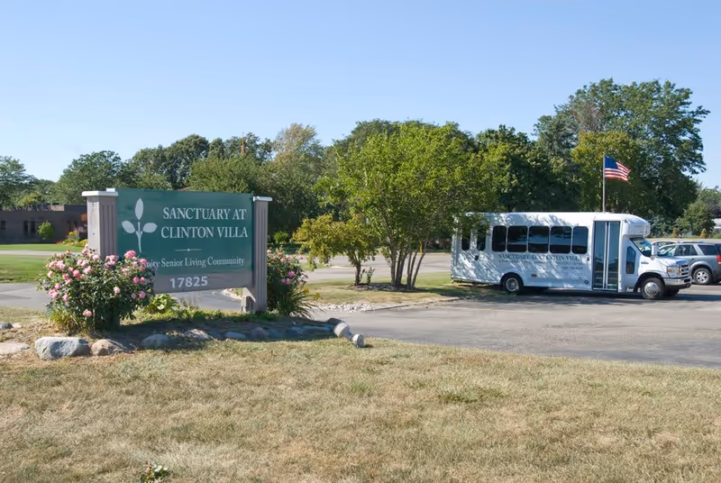 Outdoor view of the Sanctuary at Clinton Villa senior living community sign surrounded by flowers and greenery, with a white shuttle bus and parked cars in the background under a clear blue sky.