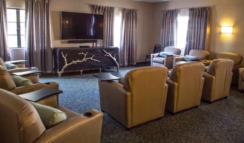 A cozy senior living common area with multiple beige recliner chairs arranged in rows facing a large flat-screen TV mounted on the wall. The room has carpeted flooring, two windows with curtains, and a unique TV stand featuring decorative branches. Soft lighting is provided by a wall-mounted lamp.