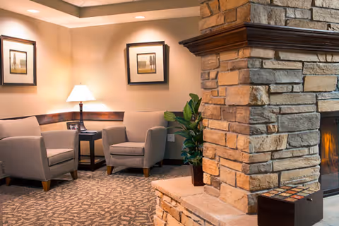 A cozy seating area with two beige armchairs and a small dark wooden side table with a lamp on it, placed against a beige wall with two framed pictures. To the right, there is a stone fireplace with a fire burning inside and a potted plant next to it. The floor is carpeted with a patterned design.