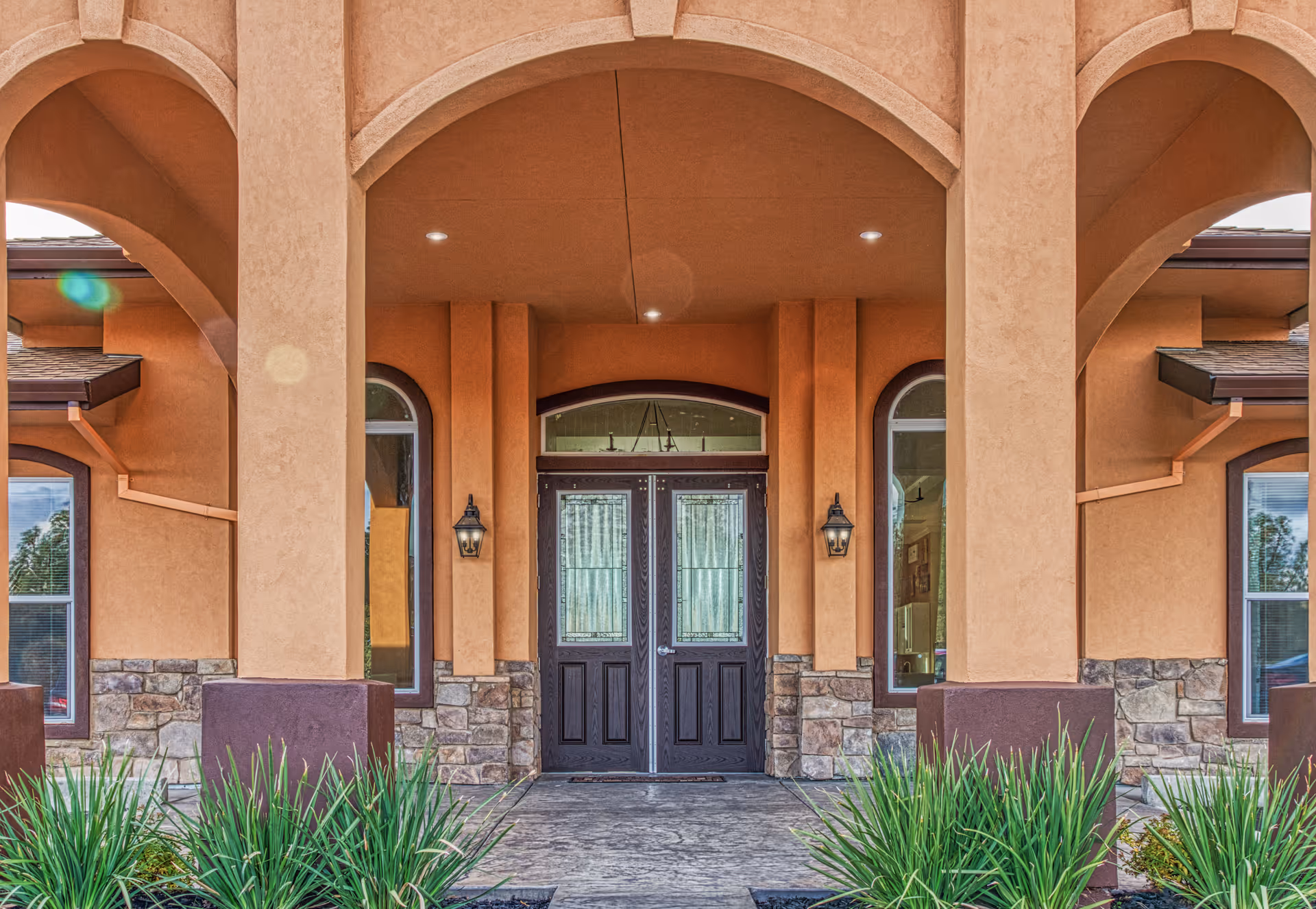 Front entrance of a senior living facility with double dark wooden doors, flanked by tall arched windows and stone accents on the lower walls. The entrance is covered by an archway with beige stucco walls and outdoor wall lanterns on either side of the doors. Green plants are visible in the foreground.