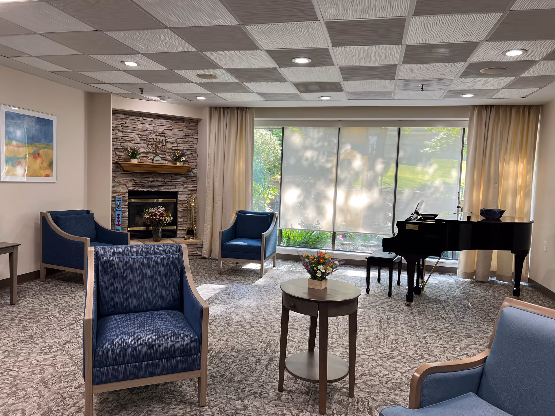 A cozy living room area with blue upholstered chairs arranged around a small round wooden table with a flower arrangement. In the background, there is a stone fireplace with decorative items on the mantel, large windows with curtains letting in natural light, and a black grand piano with a bench.