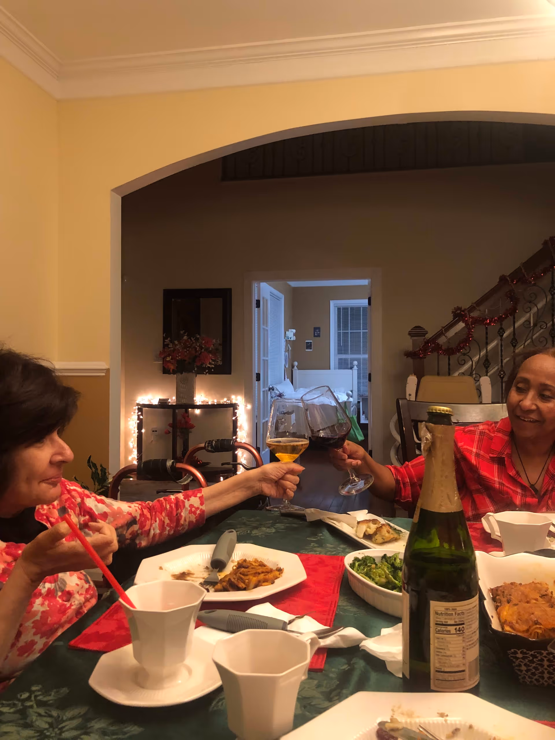 Two women sitting at a dining table in a warmly lit room, clinking wine glasses in a toast. The table is set with plates of food, cups, and a bottle of sparkling beverage. In the background, there is a doorway leading to a bedroom and a staircase decorated with red garland.