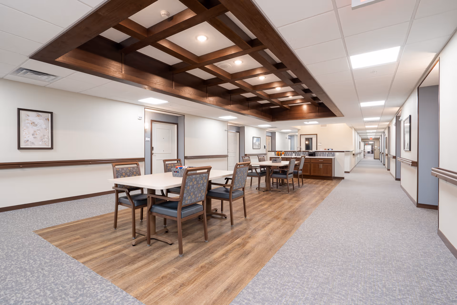 A spacious and well-lit hallway in a senior living facility with a seating area featuring two tables and several chairs on a wooden floor section. The ceiling above the seating area has a decorative wooden grid with recessed lighting. The hallway has light-colored walls with framed artwork and handrails on both sides, and doors leading to rooms along the corridor.