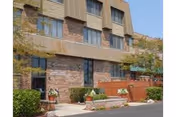 Exterior view of Briar Place Nursing Center showing a brick and beige building with multiple windows, a small landscaped area with shrubs and flowers, and a paved driveway or parking area in front.