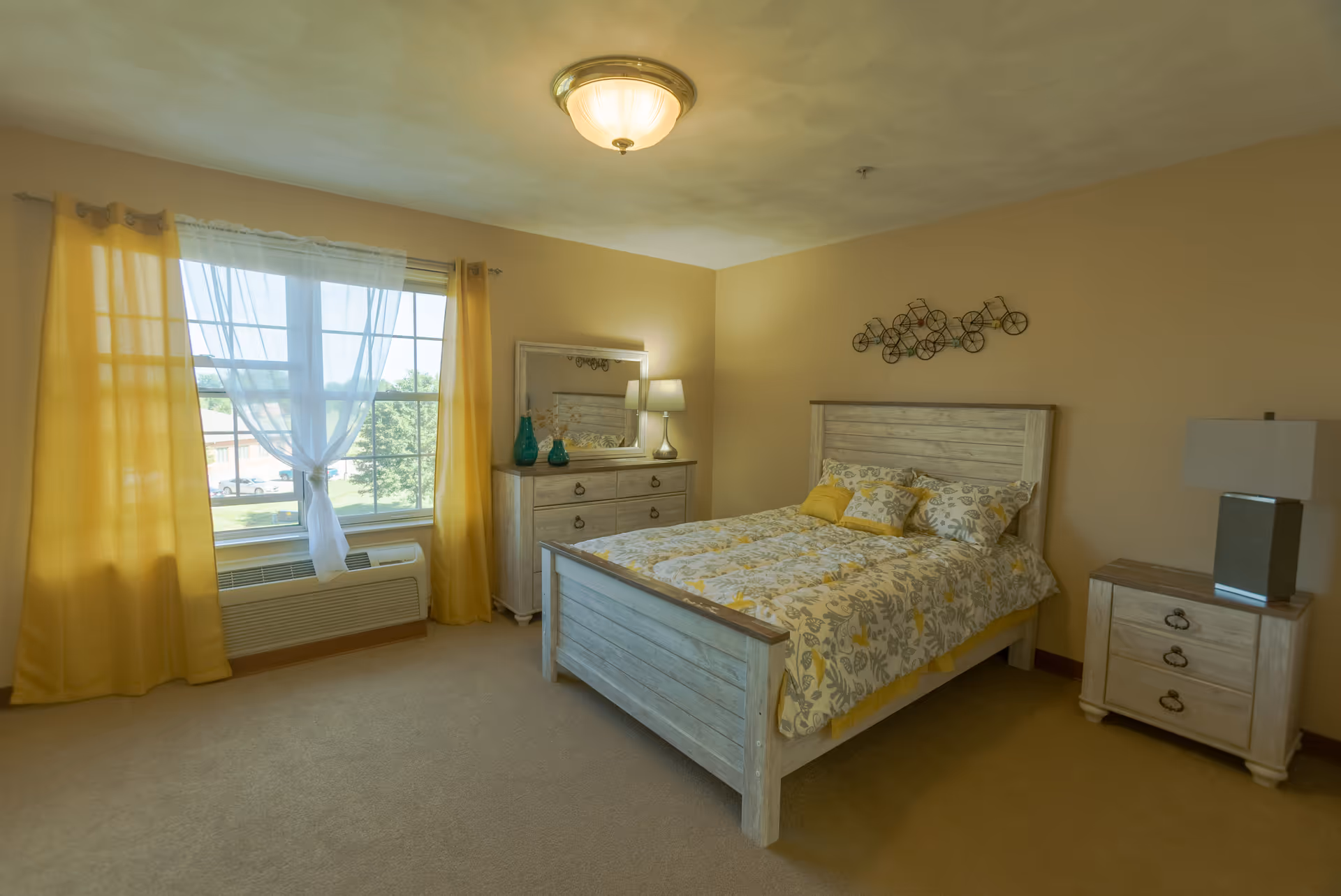 A cozy bedroom with a wooden bed frame and floral bedding in shades of yellow and gray. There is a matching wooden dresser with a mirror and two blue vases on top, a bedside table with a lamp, and a window with yellow curtains and sheer white drapes letting in natural light.