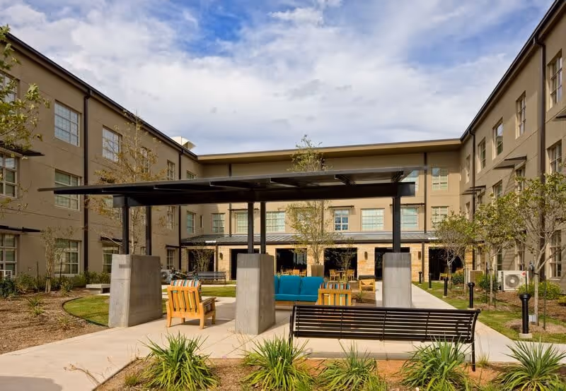 Outdoor courtyard area of a senior living facility with seating arrangements including wooden chairs, a bench, and a blue cushioned sofa under a black pergola structure. The courtyard is surrounded by a three-story building with multiple windows and landscaped greenery.