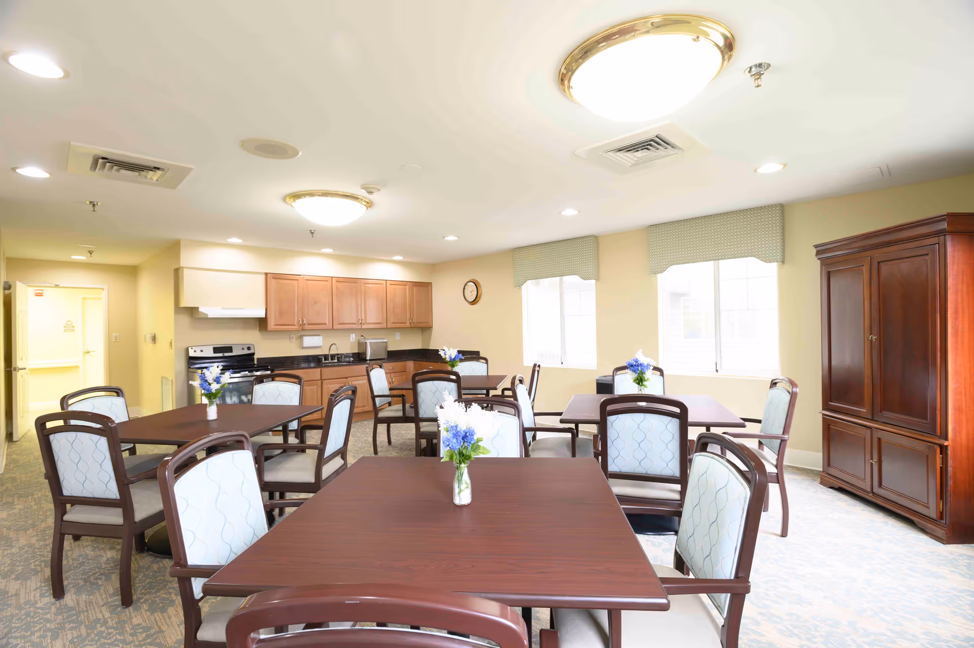 Communal dining room with several wooden tables and upholstered chairs, a kitchenette with cabinets and appliances along the back wall, and windows letting in natural light.